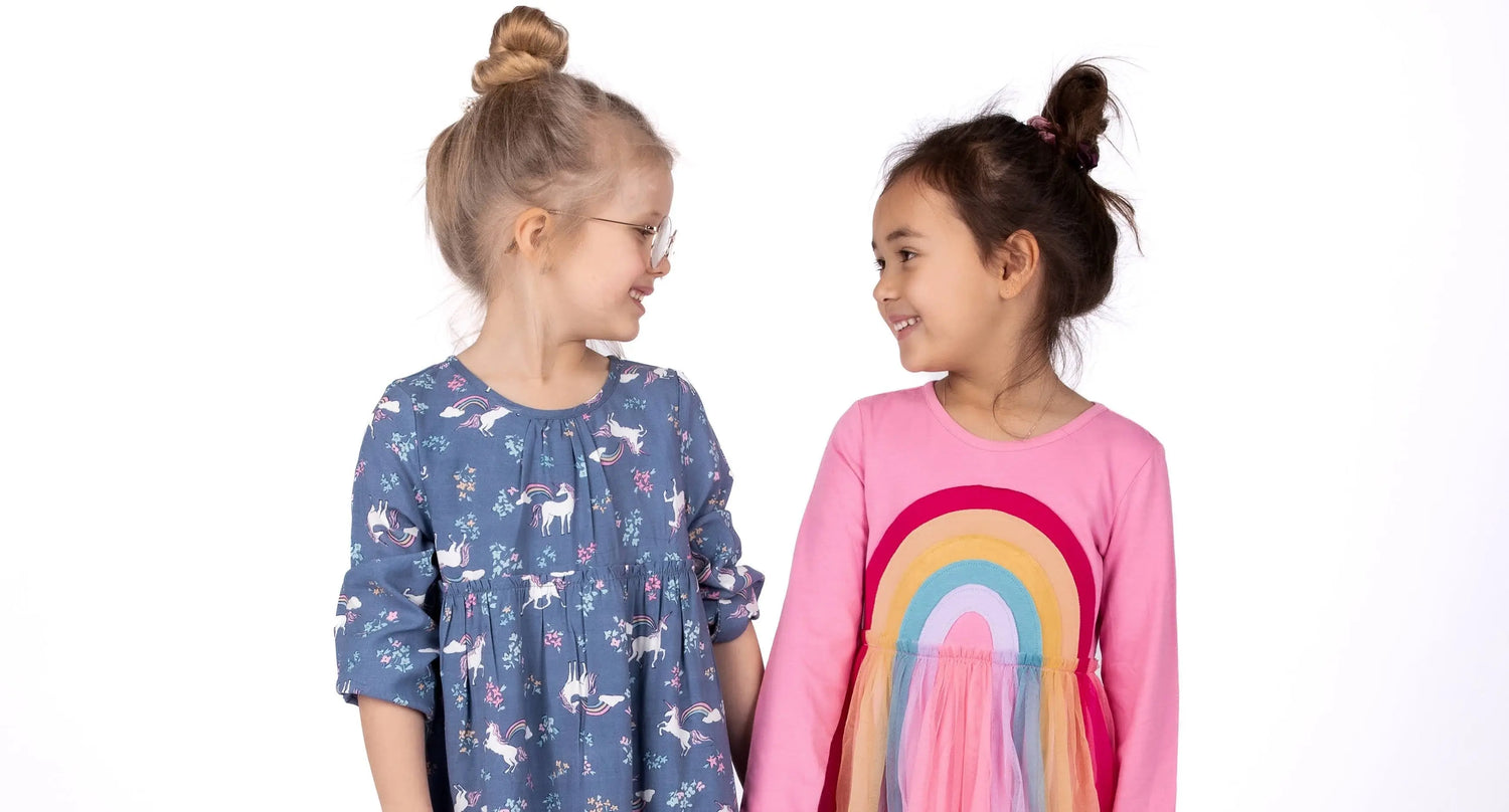 Zwei Mädchen in farbenfrohen Kleidern der Marke Danamade, lächelnd und sich ansehend, präsentiert im Mokkini Kindermode Online-Shop