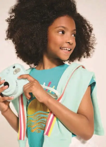 Mädchen in farbenfroher Kleidung der Marke Hatley, hält eine Kamera und lächelt, präsentiert im Mokkini Kindermode Online-Shop.