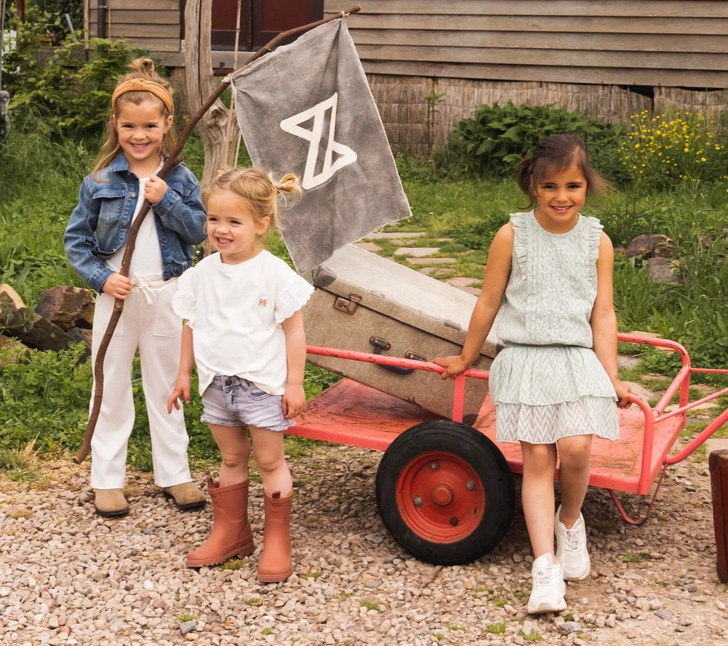 Kinder in Koko Noko Kindermode mit modernem Design – Mädchen und Junge posieren spielerisch vor einem roten Handwagen mit Flagge, umgeben von natürlicher Kulisse