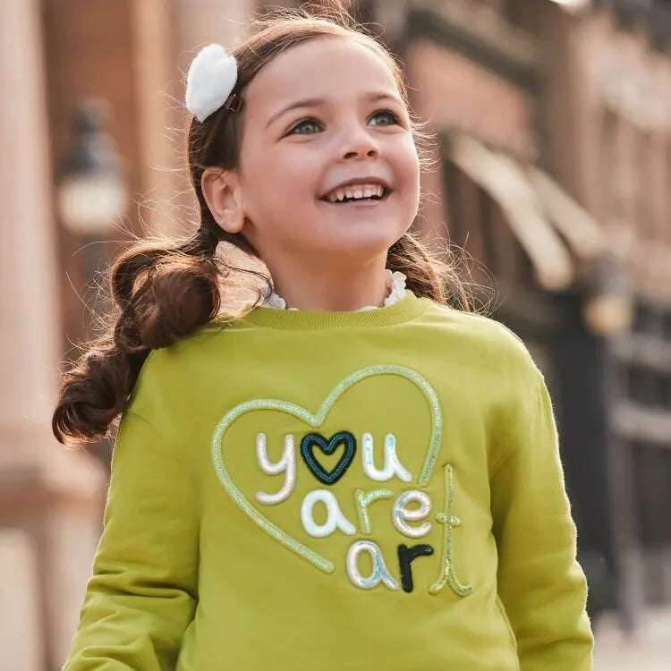 Mädchen in einem grünen Pullover mit der Aufschrift 'You Are Art' der Marke Mayoral, lächelnd im Freien, präsentiert im Mokkini Kindermode Online-Shop