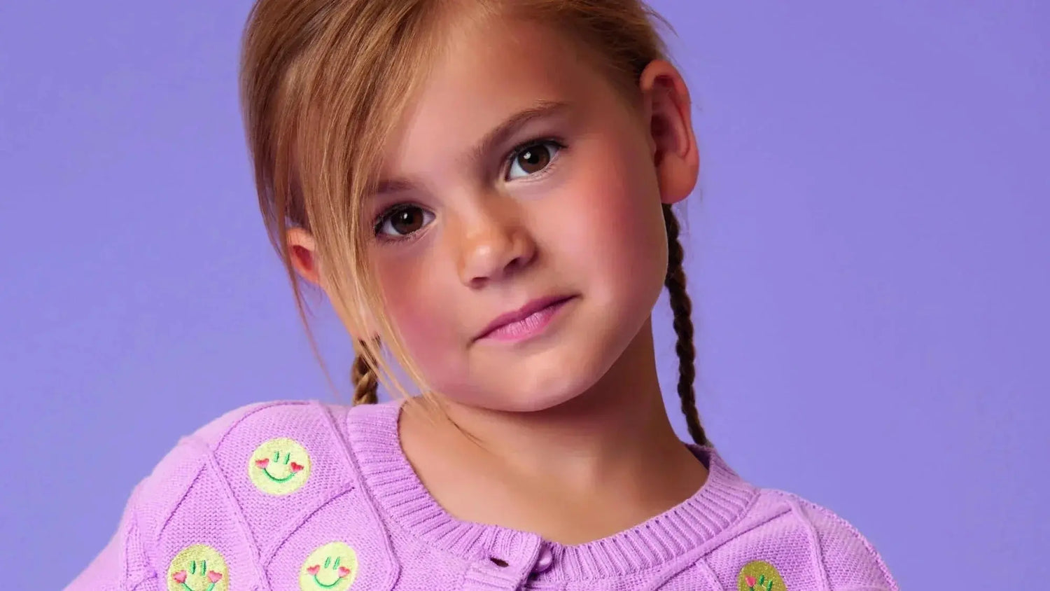"Mädchen in einem lila Strickoberteil mit Smiley-Details der Marke Oilily, vor einem lilafarbenen Hintergrund, präsentiert im Mokkini Kindermode Online-Shop