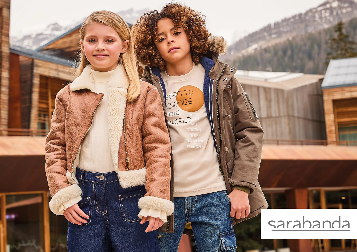 Zwei Kinder in modischer Winterkleidung der Marke Sarabanda, stehend vor einem Holzgebäude in den Bergen, präsentiert im Mokkini Kindermode Online-Shop